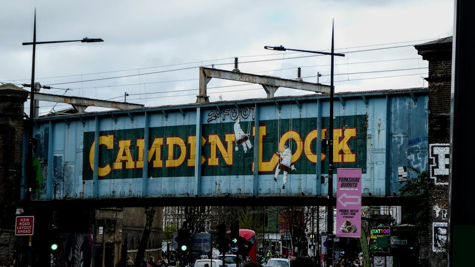 A large metal railway bridge painted in light blue, displaying the partially visible yellow and red text 'CAMDEN LOCK' in vintage-style lettering. The bridge appears weathered with some rust and peeling paint. Two graffiti-style illustrations of basketball players are painted on either side of the text, depicting figures in mid-air basketball action. Supporting the bridge are metal beams and concrete structures, with overhead train tracks and electrical lines visible against a cloudy sky background. Surrounding the scene are urban elements including streetlights, utility poles, and signage; a pink vertical banner advertising 'Yorkshire Burrito' is attached to a pole, and a small red sign indicating 'New Road Layout Ahead' is partially visible on the left side of the image. The area below the bridge shows a busy street with cars and bicycles, along with some trees that add greenery to the urban environment. This snapshot captures a typical scene in Camden, emphasizing its distinctive railway infrastructure and vibrant street atmosphere, relevant to alternative waste handling and urban clearance services such as those offered by Rubbish Clearance Camden.