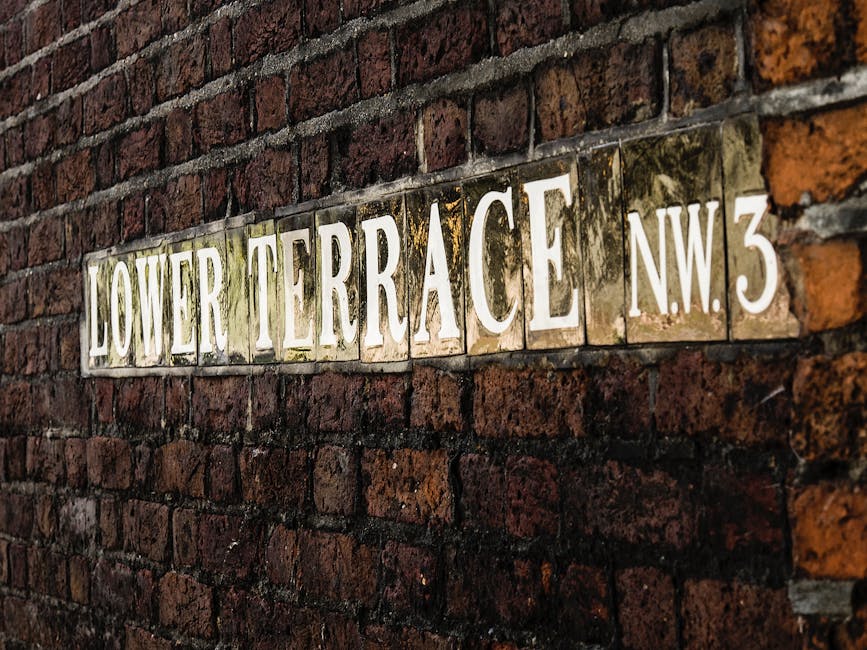 A close-up photograph of a rectangular street sign mounted on a weathered red brick wall. The sign reads 'LOWER TERRACE NW3' in large, white, capital letters with a slight glossy finish, mounted on individual ceramic tiles that have been grouted in place. The tiles have a slightly aged appearance, with some showing signs of moss or dirt buildup, especially around the edges and in the grooves. The brick wall behind the sign consists of irregularly sized, dark reddish-brown bricks with rough textures, and the mortar between them appears slightly weathered. The lighting suggests natural daylight, creating subtle shadows and highlighting the textured surfaces of both the sign and the brick wall. This scene is typical of an urban environment, possibly near a residential or commercial building, and reflects the type of infrastructure often associated with private or independent waste collection in alleyways or service areas. Rubbish Clearance Camden occasionally handles on-site clearance needs in such settings, supporting alternative waste handling methods distinct from local authority rubbish collection.