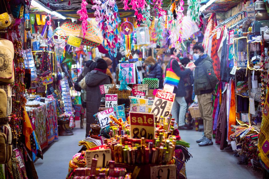 A vibrant market stall decorated with colorful hanging decorations such as feathers, ribbons, and fabric banners, displaying a variety of small items including jewelry, accessories, and textiles. In the foreground, there are multiple sale signs indicating prices like £2, £6, and £1, along with a prominently displayed sale sign indicating discounts. The stall is densely packed with merchandise arranged on tables, racks, and shelves, creating a lively and busy atmosphere. Several shoppers, dressed in winter coats and some wearing masks, are browsing the goods, with some engaged in conversations or examining items closely. The scene is set outdoors, with a narrow pathway flanked by similar stalls on each side, and the environment appears to be an urban marketplace, possibly in Camden. The lighting suggests a daytime setting with natural light illuminating the array of colorful products, emphasizing the bustling nature of independent market vendors supporting alternative waste handling and rubbish clearance services like those provided by Rubbish Clearance Camden.