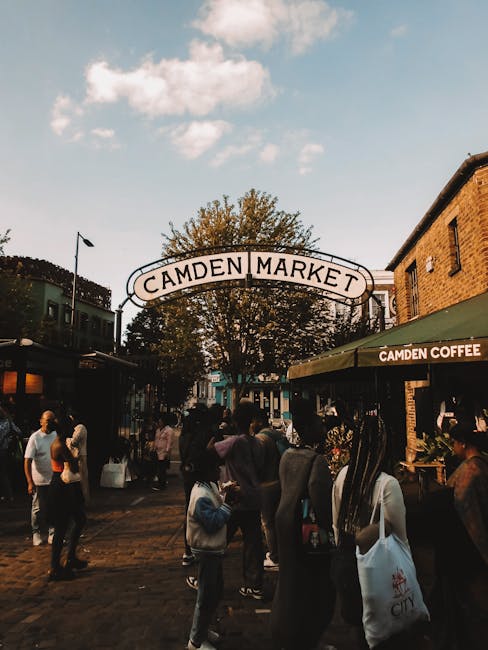 A busy scene at the entrance of Camden Market, featuring a prominent white arched sign with black lettering that reads 'Camden Market' positioned behind a large tree with dense green foliage. The market is situated in an outdoor pedestrian area paved with cobblestones, surrounded by brick-built buildings with shopfronts, including a café with a green awning displaying the words 'Camden Coffee.' Several people of varying ages and attire are walking and standing in groups, some holding shopping bags or engaging in conversations. The crowd appears lively against a background of a blue sky dotted with a few clouds, with sunlight casting gentle shadows on the scene. The setting suggests an active marketplace environment where independent vendors and local shops operate, and the scene subtly reflects an alternative approach to waste handling through informal clearing while focusing on the visual of the bustling market entrance.