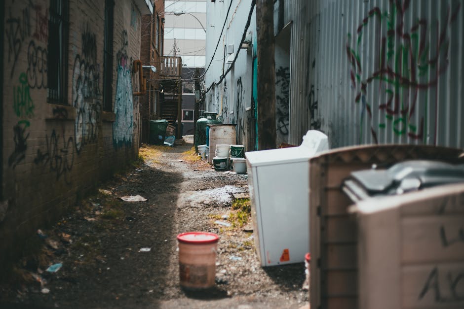 A narrow, outdoor alleyway flanked by brick and metal-clad walls, both covered in various colourful graffiti tags and street art. The ground is uneven and muddy, with patches of dirt and small puddles scattered along the pathway. On the right side, there is a large white plastic container partly visible, with a cardboard box or bin nearby; further along, two large green wheeled bins are positioned against a building. On the left, there is an orange bucket sitting on the muddy ground, with some scattered paper and debris around it. In the distance, a wooden staircase with aged, weathered steps leads to a second-floor entrance on a building at the far end of the alley. Overhead, an array of electric cables crisscross above, and ambient daylight illuminates the scene, emphasizing the gritty, urban environment typical of site-specific rubbish and waste management situations. This scene reflects an informal or alternative waste handling area, potentially serviced by independent rubbish clearance companies like Rubbish Clearance Camden for discreet rubbish removal or private waste disposal services in the area.